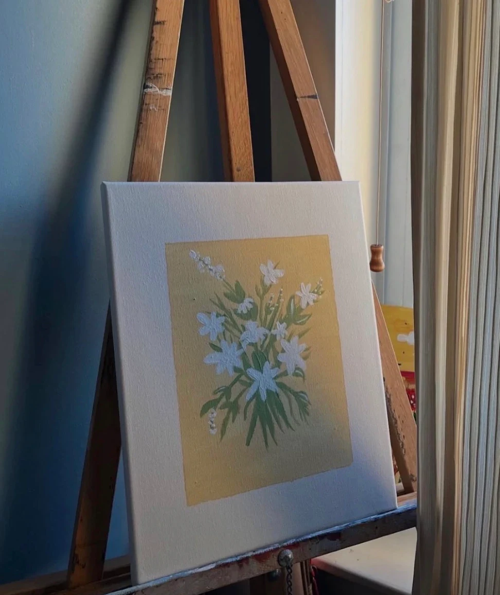 Painting of white flowers with pale green stems on a soft yellow square, on a canvas propped on a wooden easel.